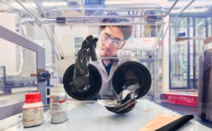 Sebuah laboratorium dengan seseorang mengenakan jas laboratorium dan sarung tangan, memegang cawan bening berisi sampel. Berbagai peralatan dan wadah laboratorium terlihat di atas meja, termasuk botol dan kain. Latar belakang menunjukkan lingkungan laboratorium modern dengan panel kaca dan peralatan.